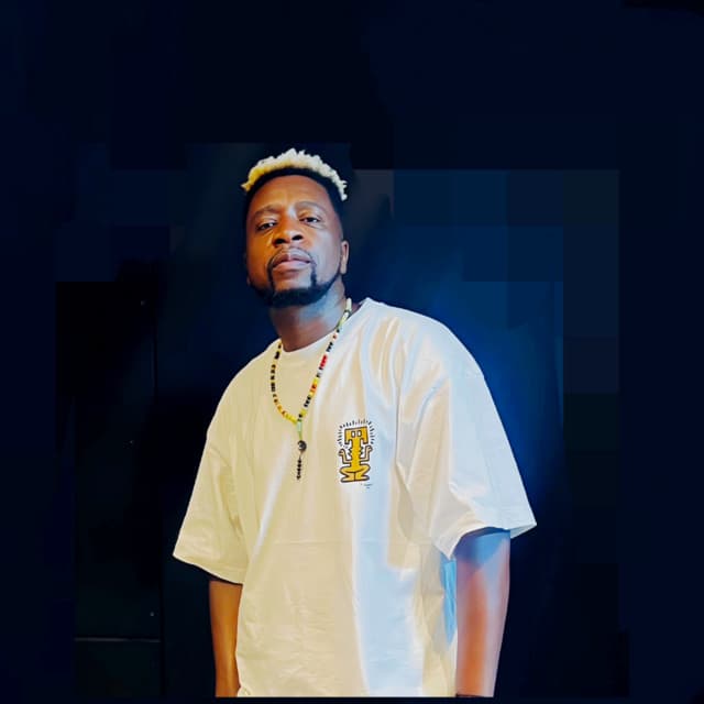 Dr Feel in a white tee and beads against a deep navy backdrop